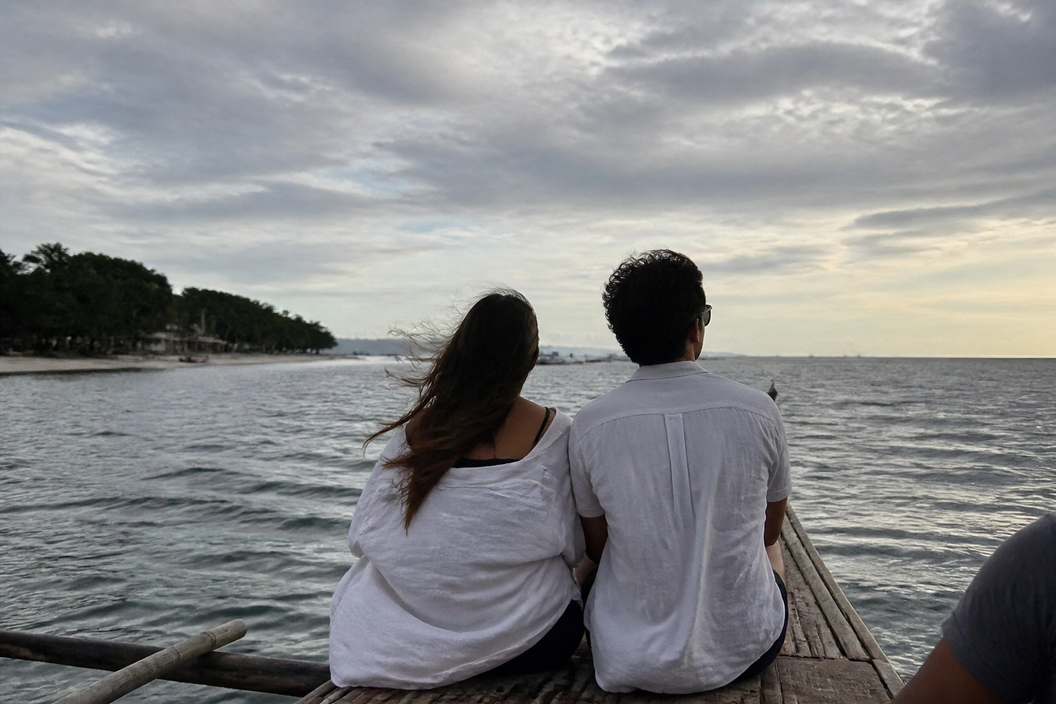 A couple sitting side by side on the bow of an outrigger boat under a cloudy evening sky
