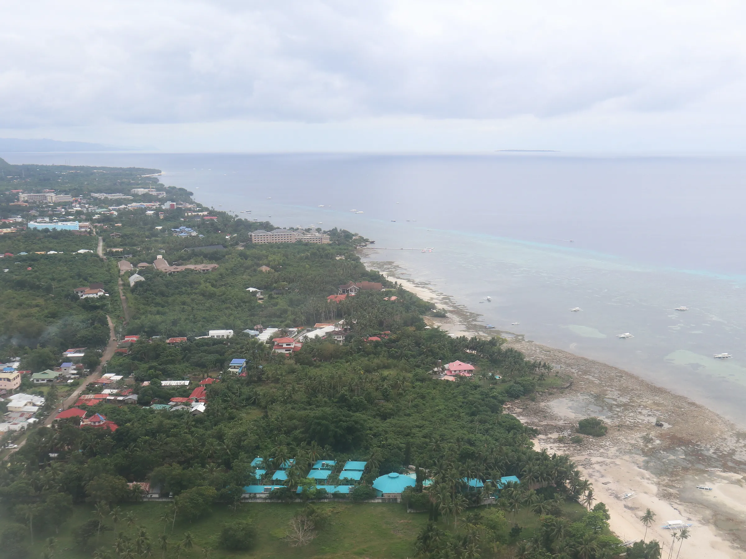 Aerial view of the Alona coast on Panglao Island: resort roofs and trees behind a pale strip of sand and shallow water